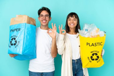 Mavi arka planda izole edilmiş plastik ve kâğıt dolu bir çanta taşıyan genç çift, parmaklarıyla 