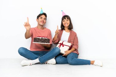 Genç melez çift, beyaz arka planda izole bir şekilde yerde oturmuş doğum gününü kutluyor ve parmağını kaldırıyor.