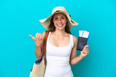 Elinde kumsal çantası ve pasaport olan beyaz bir kadın mavi arka planda izole edilmiş bir şekilde bir ürün sunmak için kenarı işaret ediyor.