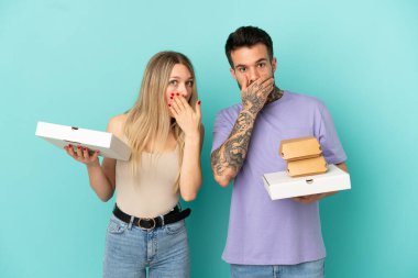 İzole edilmiş mavi arka planda pizza ve hamburger tutan çift uygunsuz bir şey söyledikleri için ağızlarını elleriyle kapatıyor.
