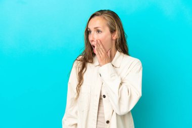 Mavi arka planda izole edilmiş genç beyaz kadın yan tarafa bakarken sürpriz bir jest yaparak bir şeyler fısıldıyor.