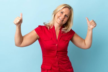 Mavi arka planda izole edilmiş orta yaşlı bir kadın baş parmak işareti yapıyor.
