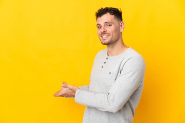 Sarı arka planda izole edilmiş beyaz tenli yakışıklı adam, davet edilmek için ellerini yana uzatıyor.