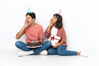 Genç ve melez bir çift, doğum gününü kutluyorlar. Beyaz arka planda izole bir şekilde oturuyorlar. Ağızları sonuna kadar açık bağırıyorlar.