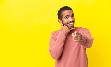 Sarı arka planda izole edilmiş genç latin adam telefon hareketi yapıyor ve ön tarafı işaret ediyor.