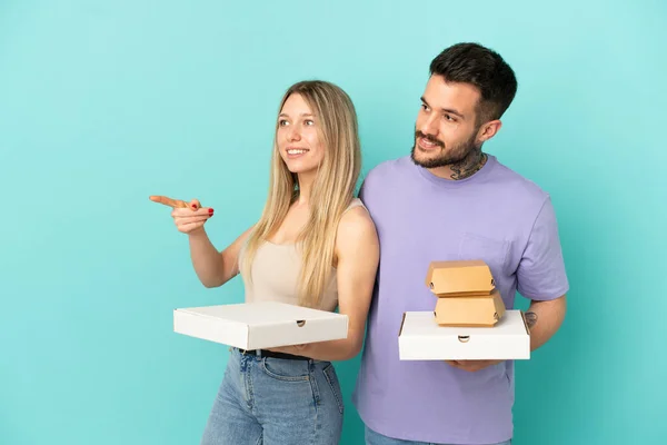 Couple holding pizzas and burgers over isolated blue background presenting an idea while looking smiling towards