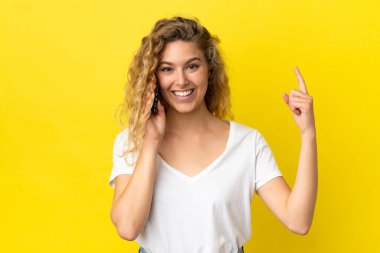 Sarı arka planda izole edilmiş cep telefonu kullanan genç sarışın kadın harika bir fikre işaret ediyor.