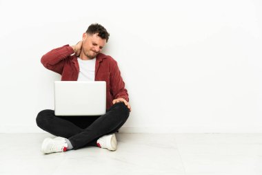 Genç, yakışıklı, beyaz bir adam dizüstü bilgisayarı ve boyun ağrısı ile yerde oturuyor.