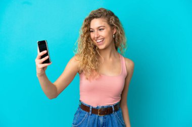 Mavi arka planda izole edilmiş genç sarışın bir kadın selfie çekiyor.