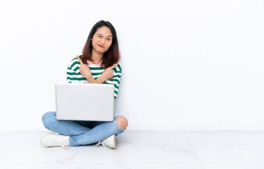 Yerde dizüstü bilgisayarı olan genç Vietnamlı bir kadın beyaz duvarda izole bir şekilde oturuyor. Yan taraftakileri işaret ediyor.