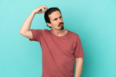 Kafkasyalı genç adam mavi arka planda izole edilmiş. Kafası kaşınırken şüpheleri var.