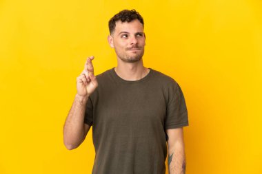 Sarı arka planda izole edilmiş beyaz, yakışıklı genç bir adam parmaklarını çapraz yapıp en iyisini diliyor.