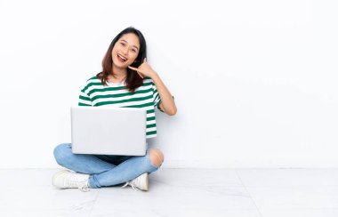 Yerde dizüstü bilgisayarı olan genç Vietnamlı bir kadın, beyaz duvarda izole bir şekilde telefon jesti yapıyor. Bana geri dönüş işareti ver.