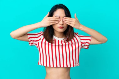 Genç Vietnamlı bir kadın mavi arka planda izole edilmiş. Gözlerini elleriyle kapatmış ve gülümsüyor.