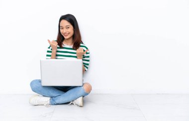 Dizüstü bilgisayarı olan genç Vietnamlı kadın beyaz duvarda izole bir şekilde oturuyor. Baş parmakları havada ve gülümsüyor.