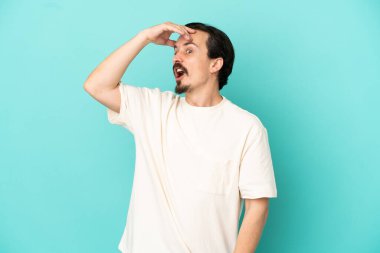 Mavi arka planda izole edilmiş genç beyaz adam yan tarafa bakarken sürpriz jest yapıyor.