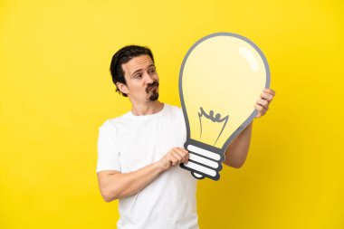 Sarı arka planda izole edilmiş genç beyaz bir adam elinde bir ampul simgesi ve şüpheleri var.