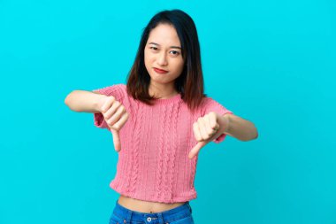 Mavi arka planda izole edilmiş genç Vietnamlı kadın baş parmağını iki eliyle aşağı gösteriyor.