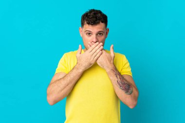 Beyaz tenli yakışıklı adam mavi arka planda izole edilmiş, ağzı elleriyle kapatıyor.