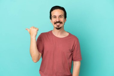 Mavi arka planda izole edilmiş genç beyaz adam bir ürünü sunmak için kenarı işaret ediyor.