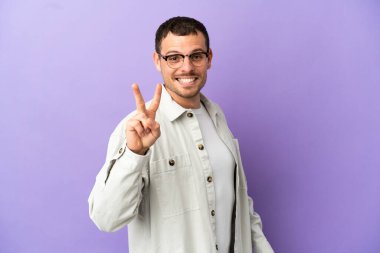 İzole edilmiş mor arka planda Brezilyalı adam gülümsüyor ve zafer işareti gösteriyor