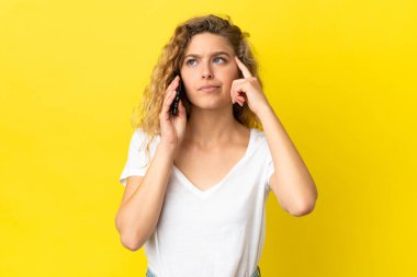 Sarı arka planda izole edilmiş cep telefonu kullanan genç sarışın kadının şüpheleri ve düşünceleri var.