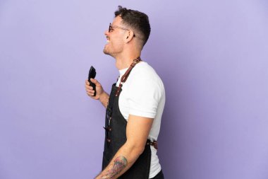 Önlüklü berber beyaz adam mor arka planda izole edilmiş. Yan pozisyonda gülüyor.