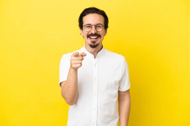 Sarı arka planda izole edilmiş genç beyaz adam şaşırmış ve ön tarafı işaret ediyor.