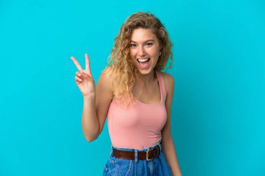 Mavi arka planda izole edilmiş genç sarışın kadın gülümsüyor ve zafer işareti gösteriyor.