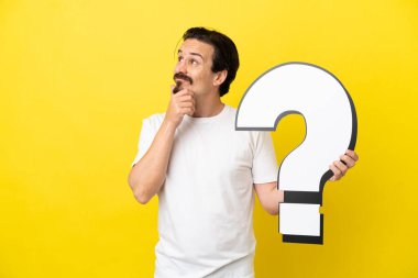 Sarı arka planda izole edilmiş genç beyaz bir adam elinde bir soru işareti simgesi tutuyor ve şüpheleri var.
