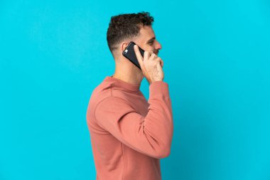 Beyaz tenli yakışıklı adam mavi arka planda izole edilmiş cep telefonuyla biriyle sohbet ediyor.