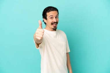 Kafkasyalı genç adam mavi arka planda izole edildi. Başparmakları havada çünkü iyi bir şey oldu.