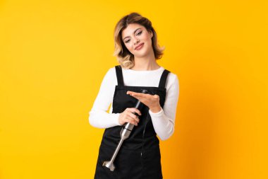 Sarı arkaplanda izole edilmiş el blenderi kullanan kız gülümseyerek bir fikir sunuyor.