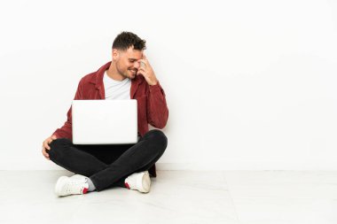 Genç, yakışıklı, beyaz bir adam yerde oturmuş dizüstü bilgisayarla gülüyor.