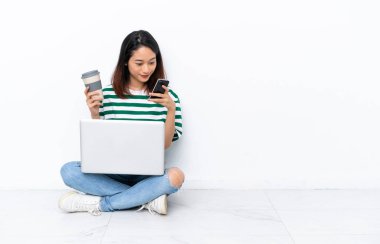 Yerde dizüstü bilgisayarı olan genç Vietnamlı bir kadın beyaz duvarda izole bir şekilde oturuyor. Elinde kahve ve cep telefonu var.