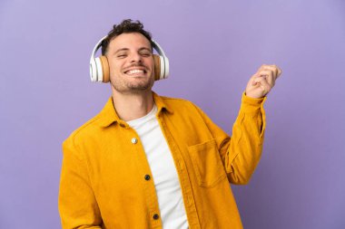 Genç, beyaz, yakışıklı bir adam mor arka planda izole edilmiş müzik dinliyor ve gitar hareketi yapıyor.