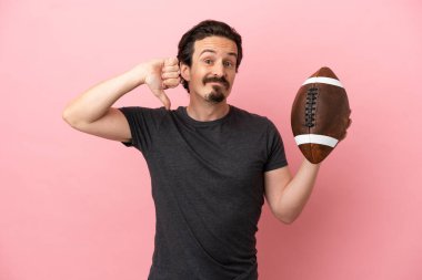 Pembe arka planda ragbi oynayan genç, beyaz bir adam, olumsuz bir ifadeyle baş parmağını aşağı gösteriyor.