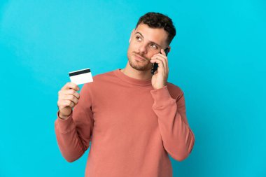 Beyaz tenli yakışıklı adam mavi arka planda izole edilmiş cep telefonuyla sohbet ediyor ve elinde bir kredi kartı tutuyor.