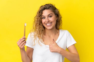 Sarı arka planda dişlerini fırçalayan beyaz bir kadın baş parmak hareketi yapıyor.