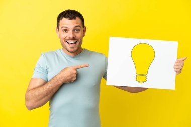 İzole edilmiş mor arka plan üzerinde Brezilyalı bir adam elinde ampul simgesi olan bir levha tutuyor ve ona doğrultuyor.