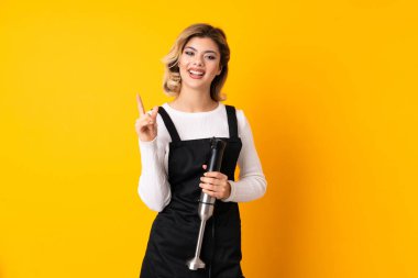 Sarı arka planda izole edilmiş el blenderi kullanan kız harika bir fikre işaret ediyor