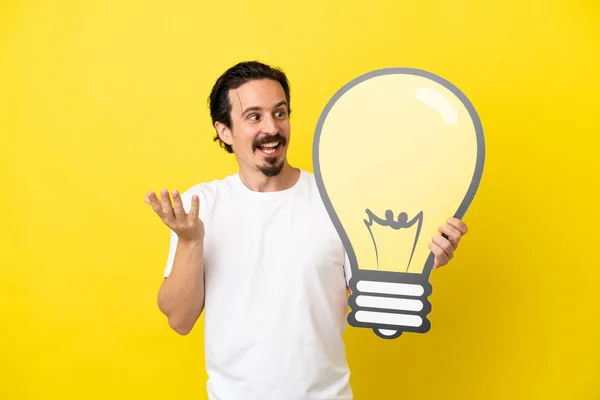Young Caucasian Man Isolated Yellow Background Holding Bulb Icon Surprised — Stock Photo, Image