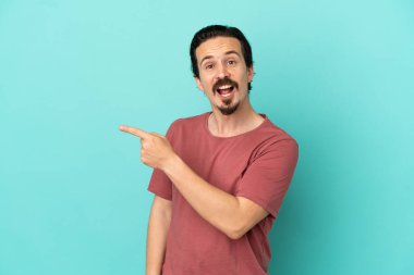 Mavi arka planda izole edilmiş genç beyaz adam şaşırmış ve işaret ediyor.