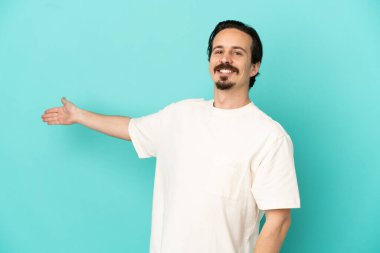 Mavi arka planda izole edilmiş genç beyaz adam, davet edilmek için ellerini yana uzatıyor.