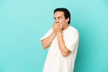 Genç beyaz adam mavi arka planda izole edilmiş ağzını kapatıyor ve yan tarafa bakıyor.