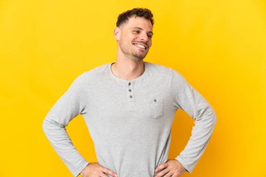 Sarı arka planda izole edilmiş, kolları kalçasında ve gülümseyen genç beyaz bir adam.