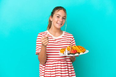 Mavi arka planda izole edilmiş waffle tutan küçük beyaz kız para hareketi yapıyor.