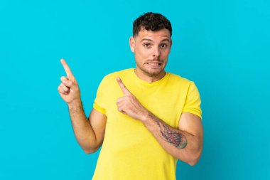 Kafkasyalı yakışıklı adam mavi arka planda izole edilmiş, korkmuş ve kenarı işaret ediyor.