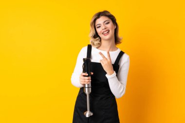 Sarı arka planda izole edilmiş el blenderi kullanan kız gülümsüyor ve zafer işareti gösteriyor