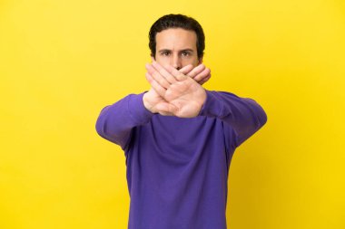 Sarı arka planda izole edilmiş genç beyaz bir adam bir eylemi durdurmak için eliyle jesti durduruyor.